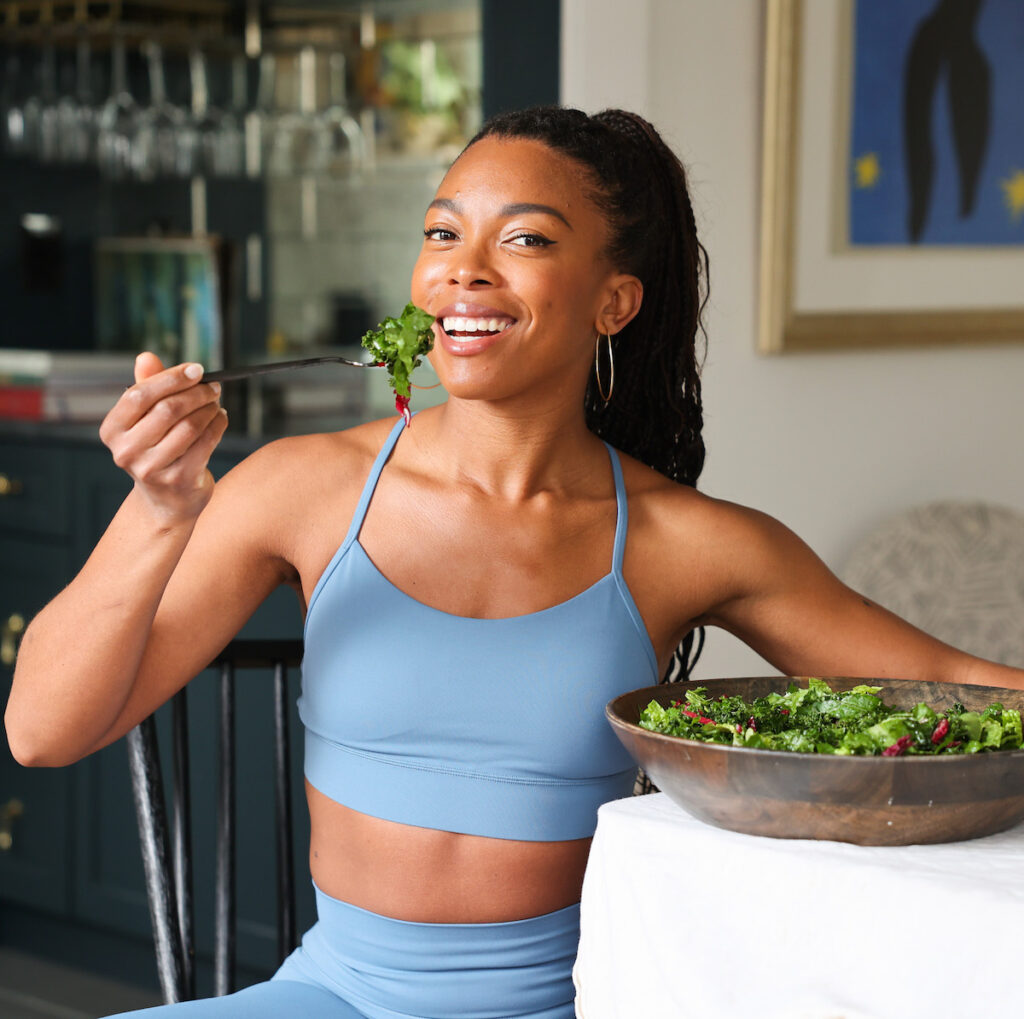 Jenne Claiborne eating a large salad at table wearing blue activewear