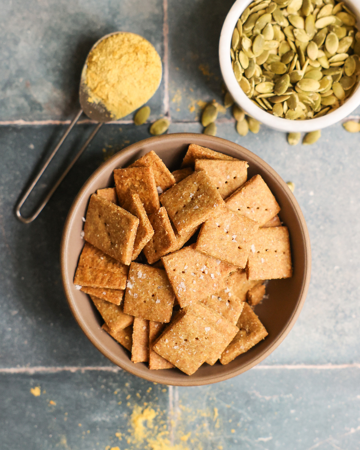 An overhead shot of a bowl of pumpkin seed crackers on the counter with pumpkin seeds and a spoonful of nutritional yeast.