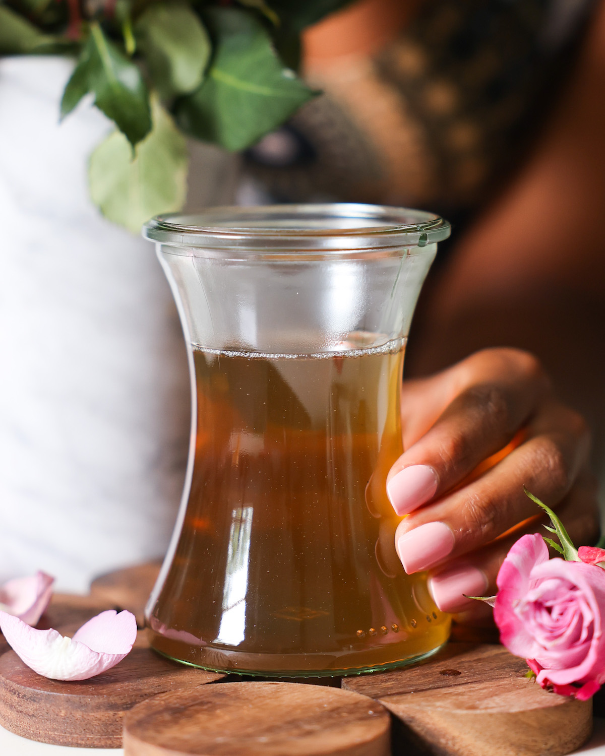 rose simple syrup in a glass jar held by a brown skinned hand, surrounded by rose petals. 