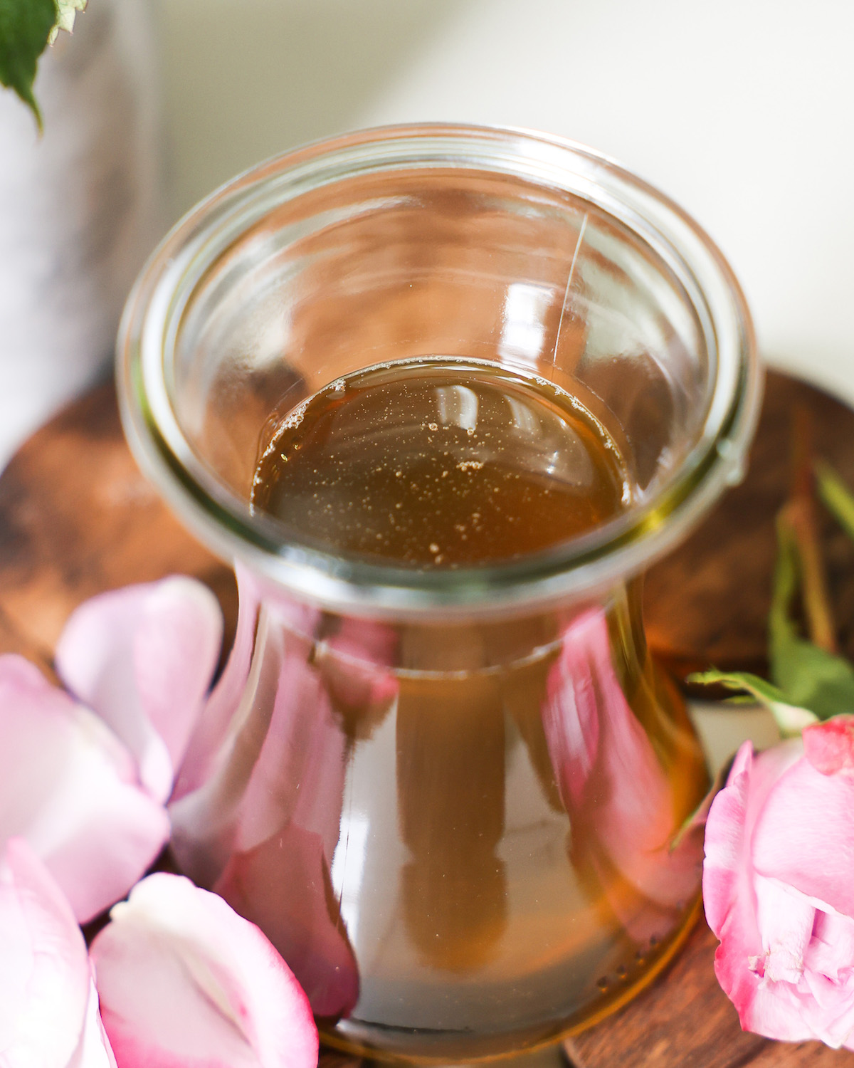 Image of homemade rose simple syrup in a glass jar surrounded by rose petals. 