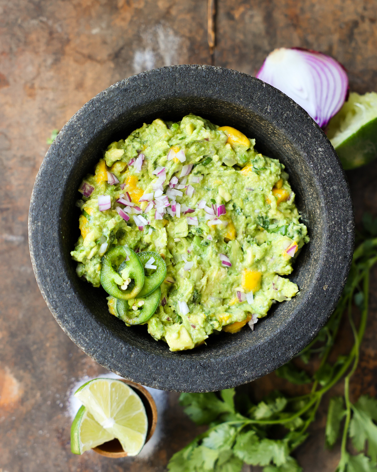 An overhead shot of mango guacamole in a mortar on the counter with red onions and limes.