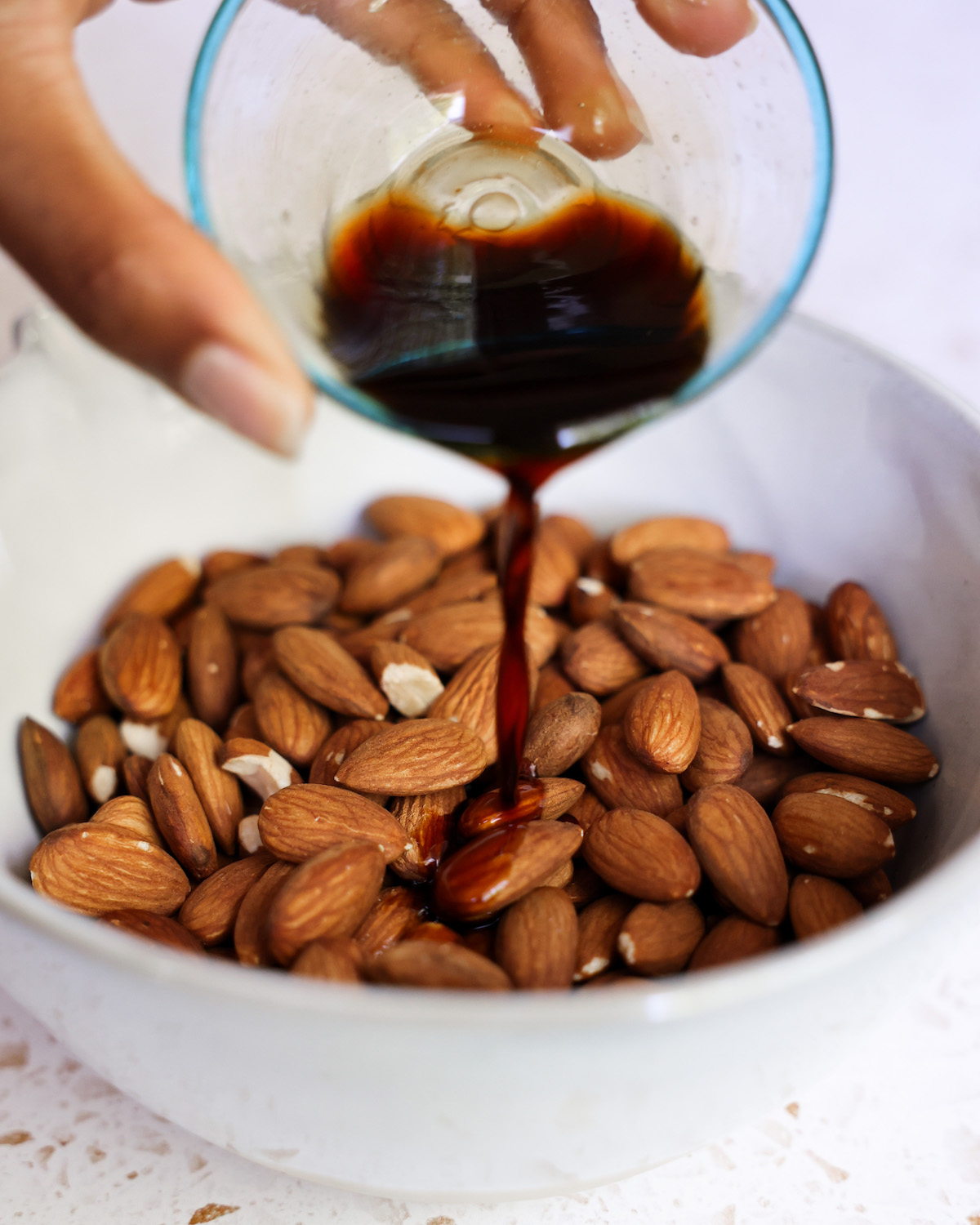 Drizzling a small dish of tamari over hot roasted almonds in a white bowl.
