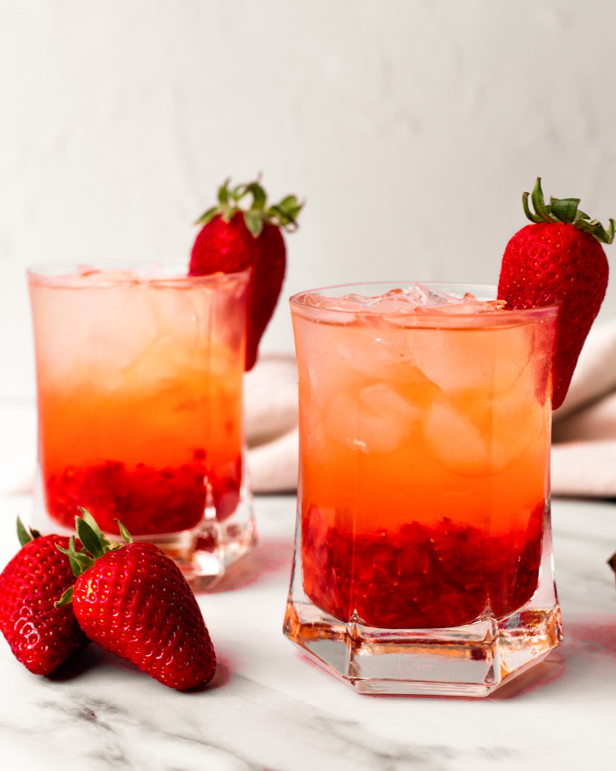 A side shot of two glasses of strawberry rose cocktail with strawberries on the side of the classes.