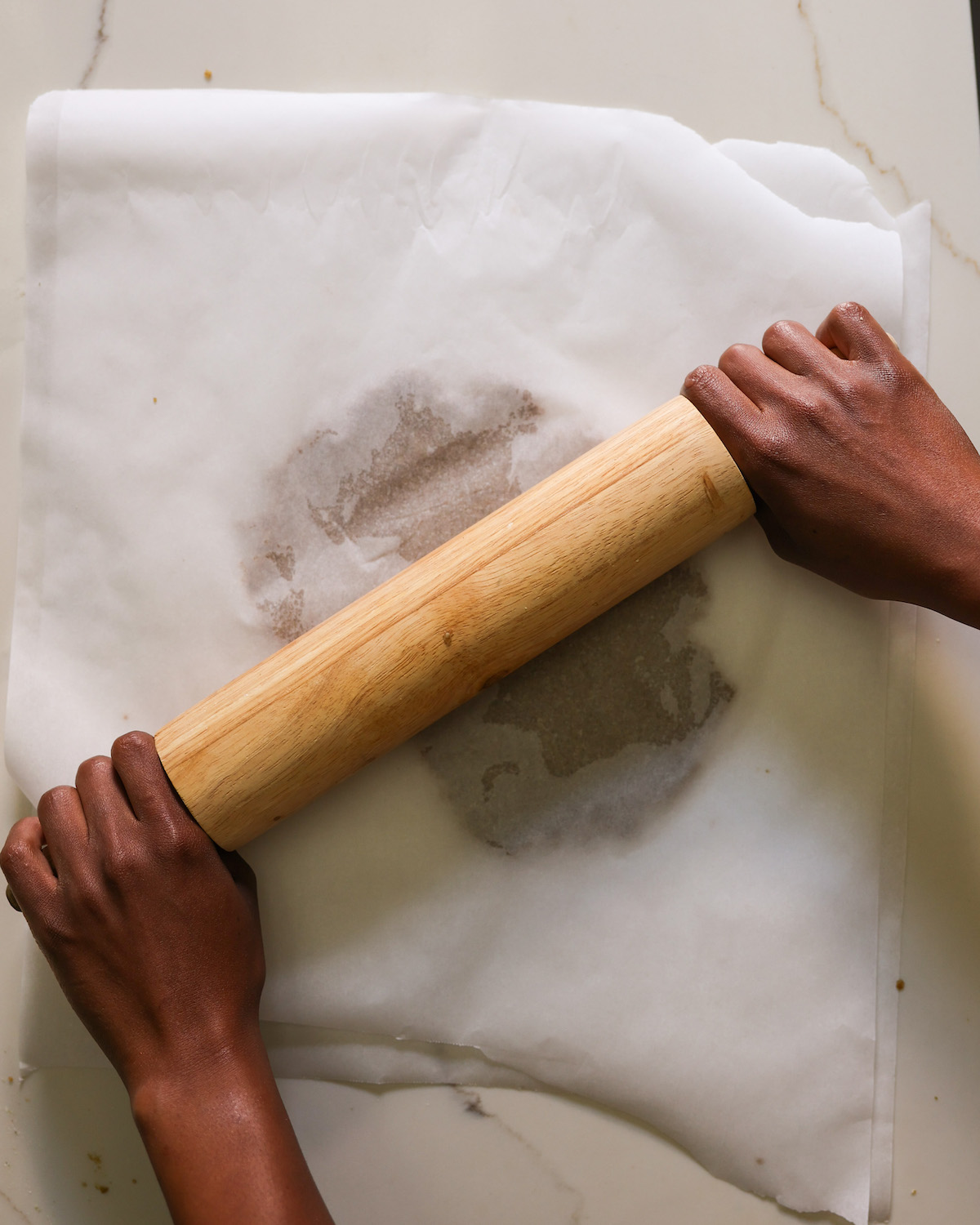 Brown hands rolling out the pumpkin seed cracker dough between two sheets of parchment paper.