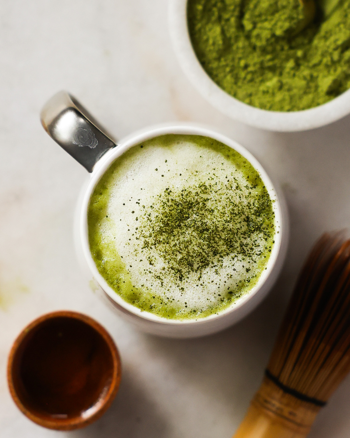 An overhead shot of a mug of the oat milk matcha latte on the counter next to a small dish of matcha and the matcha whisk.