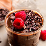 An up-close angled shot of a glass dish of vegan chocolate mousse on the counter with carob chips and raspberries.