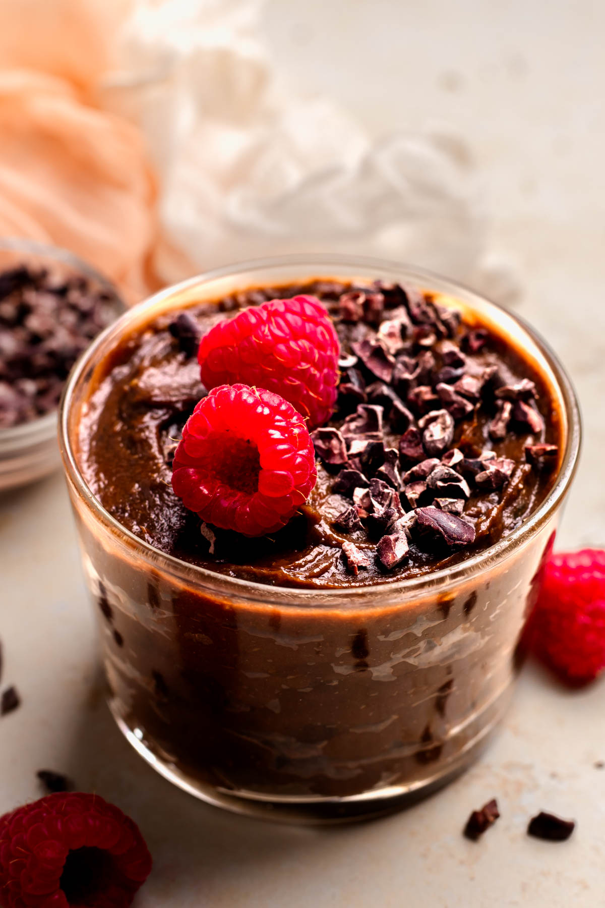 An up-close angled shot of a glass dish of vegan chocolate mousse on the counter with carob chips and raspberries.