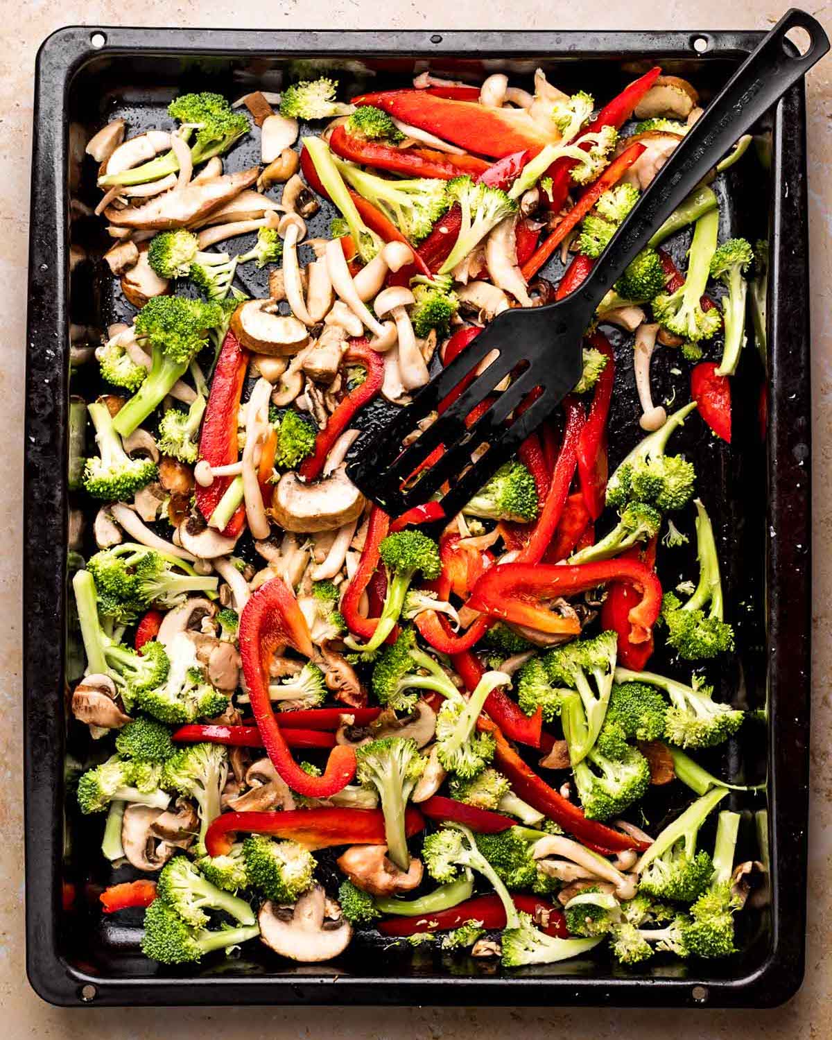 An overhead shot of a baking sheet of sliced vegetables with a black spatula.