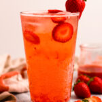 A side shot of a tall glass of strawberry soda on the counter with strawberries and a dish towel.