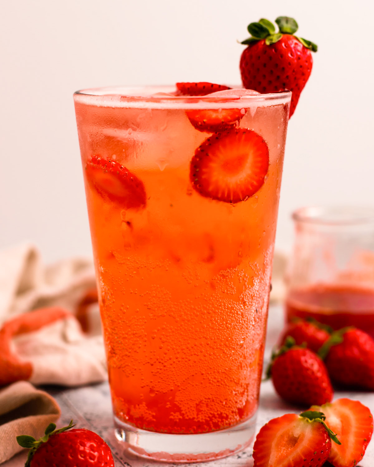 A side shot of a tall glass of strawberry soda on the counter with strawberries and a dish towel.