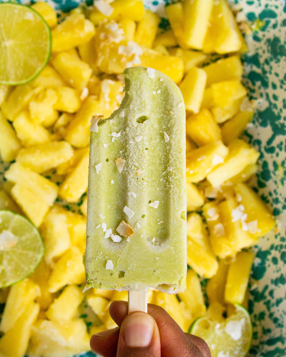 A closeup overhead shot of a brown hand holding an avocado pineapple popsicle with a bite taken out of it.