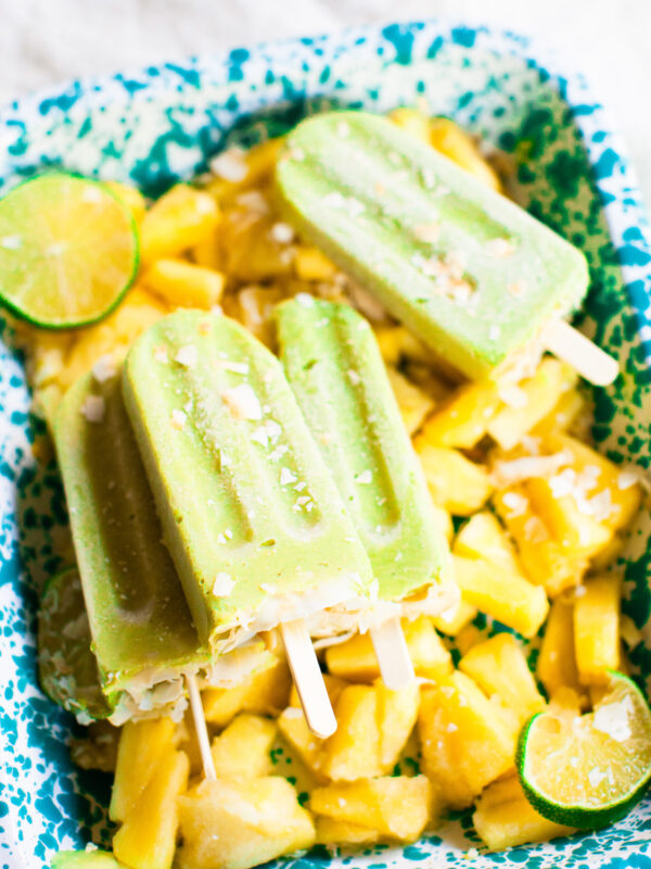 An angled side shot of avocado pineapple popsicles stacked on a blue tray of frozen pineapple.