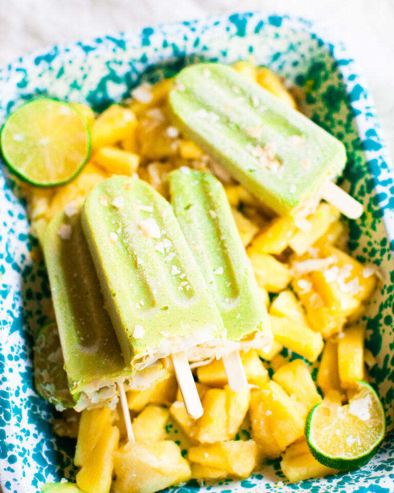 An angled side shot of avocado pineapple popsicles stacked on a blue tray of frozen pineapple.