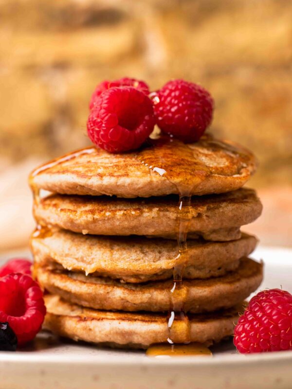 An up-close side shot of a small stack of spelt pancakes with raspberries and maple syrup.