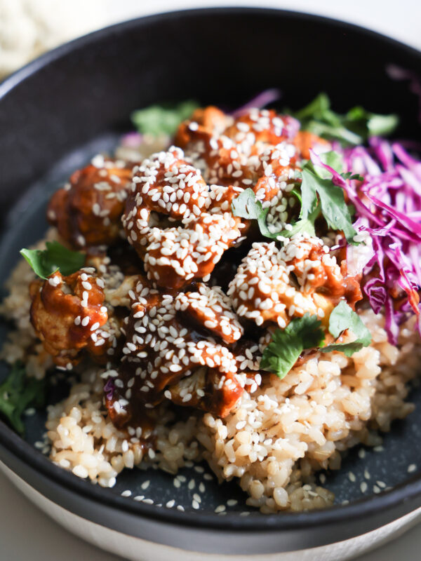 A closeup shot of the orange cauliflower with rice, red cabbage, and cilantro in a black bowl.