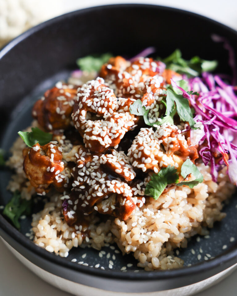 A closeup shot of the orange cauliflower with rice, red cabbage, and cilantro in a black bowl.
