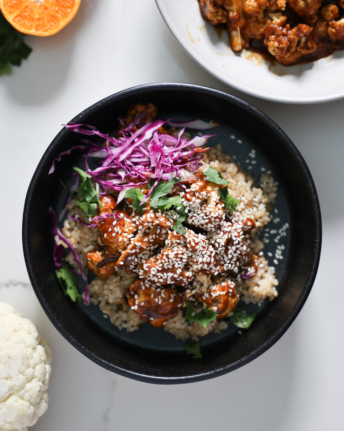 An overhead shot of a black bowl of the orange cauliflower with sesame seeds, red cabbage, and cilantro.