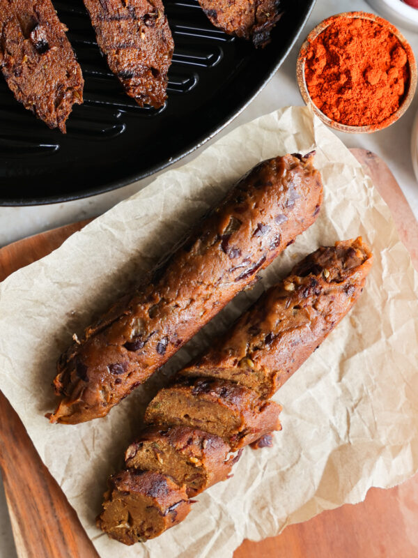 An overhead shot of two vegan sausages-- one whole and one sliced -- on a parchment paper-lined cutting board.