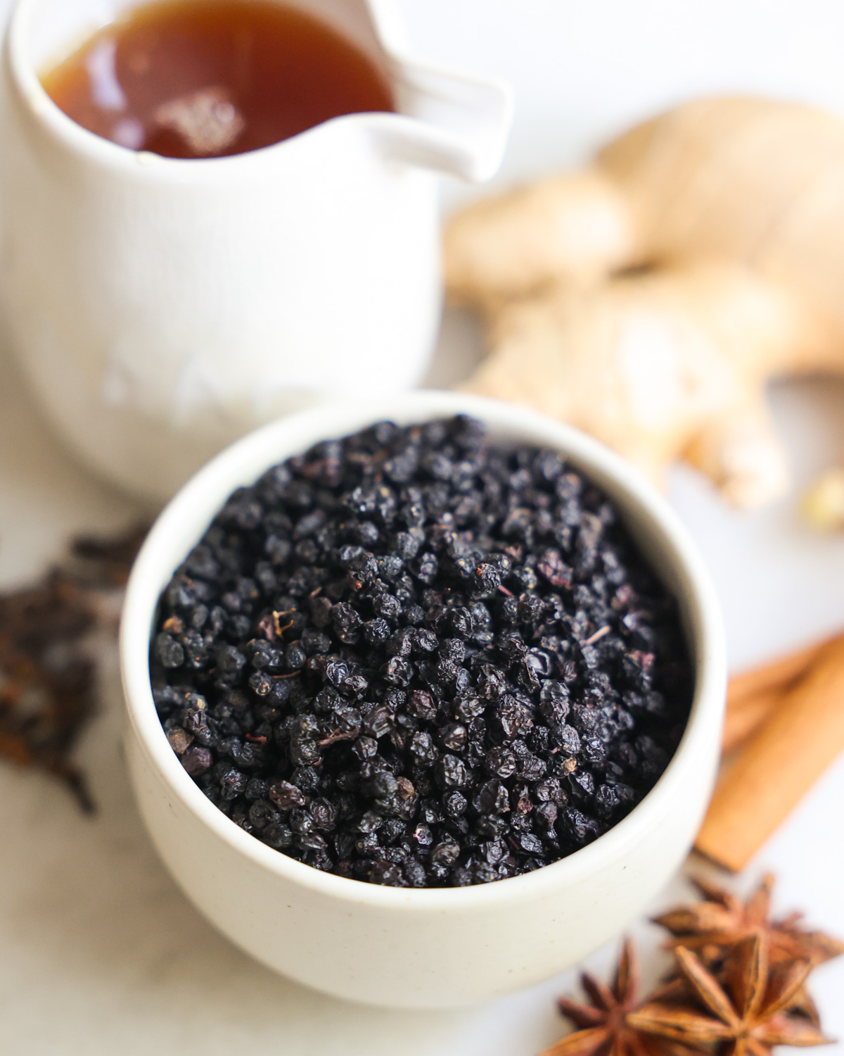 An up-close shot of dried elderberries in a small white bowl with spices and agave syrup in the background.