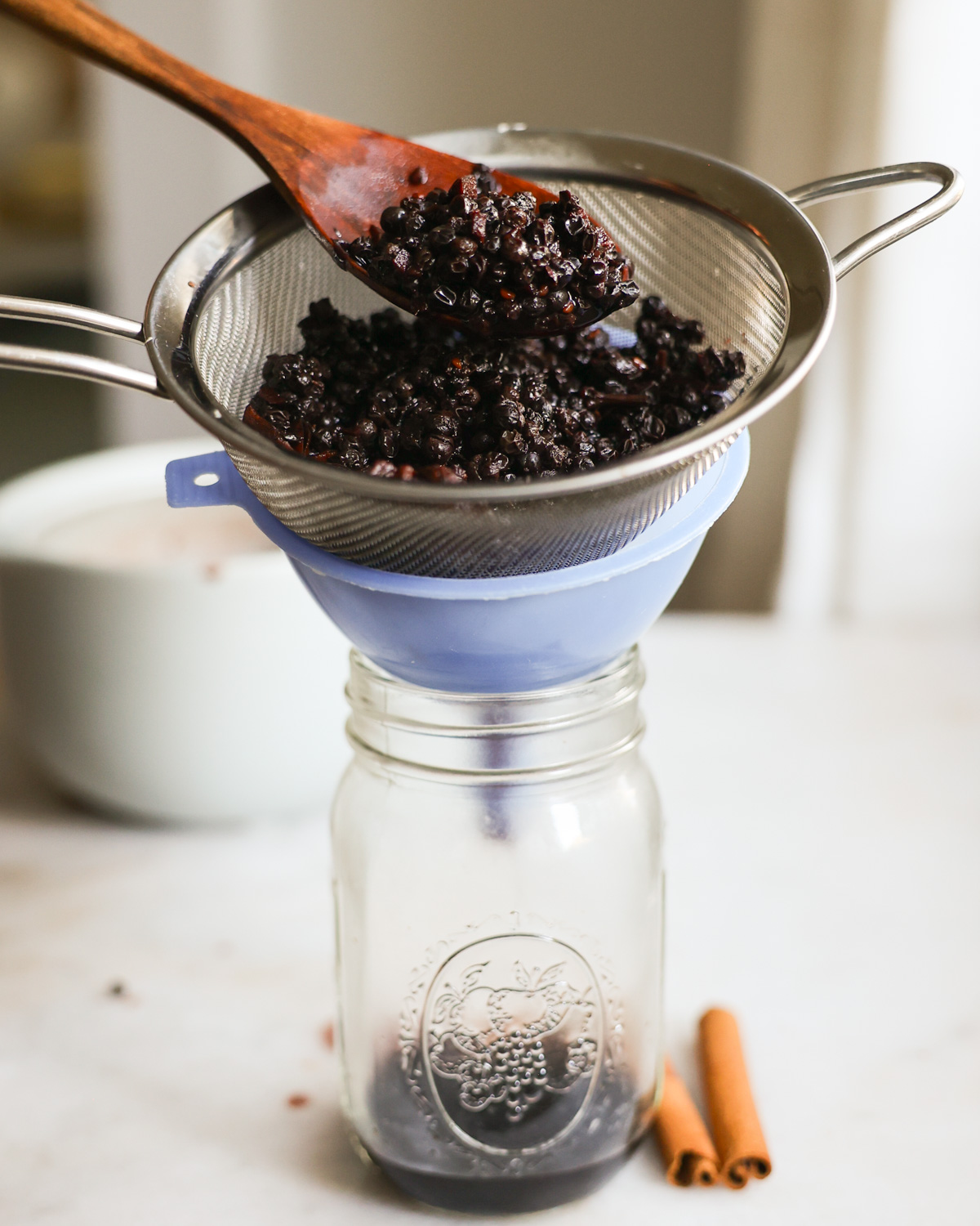 Pressing the simmered elderberry syrup through a sieve over a funnel into a glass jar.