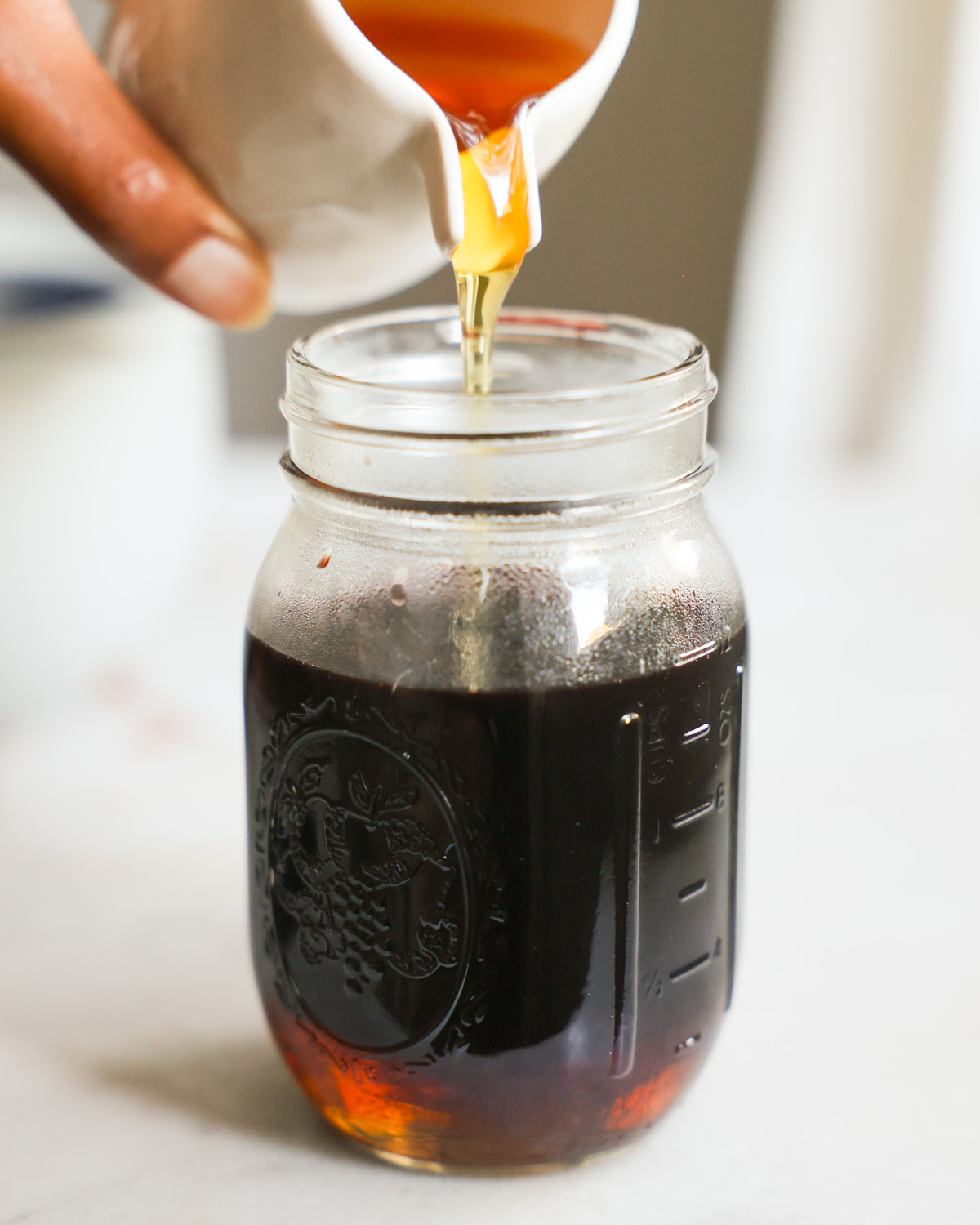 An up-close shot of a brown hand pouring agave syrup into a jar with the elderberry juice.