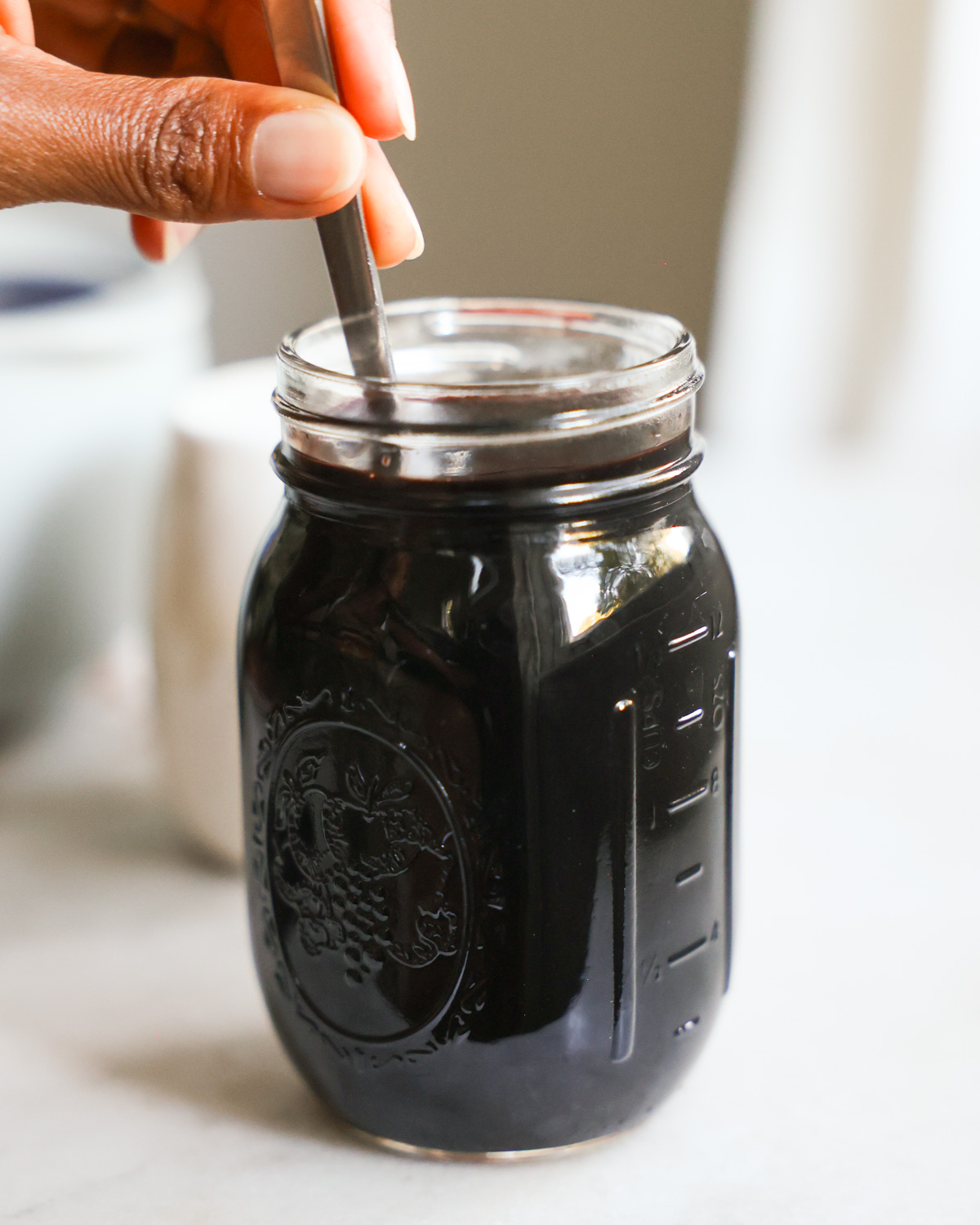 A close-up shot of a brown hand hand stirring a small har of elderberry syrup.