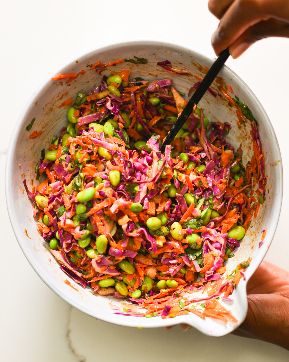 A brown hand stirring a bowl of edamame salad.