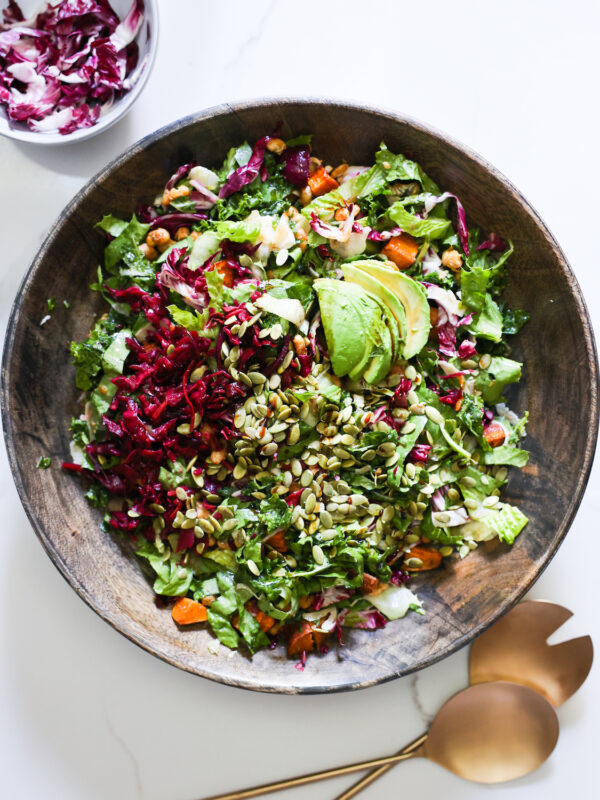 An overhead shot of the autumn kale salad in a mixing bowl with metal salad tongs.