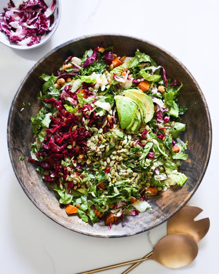 An overhead shot of the autumn kale salad in a mixing bowl with metal salad tongs.