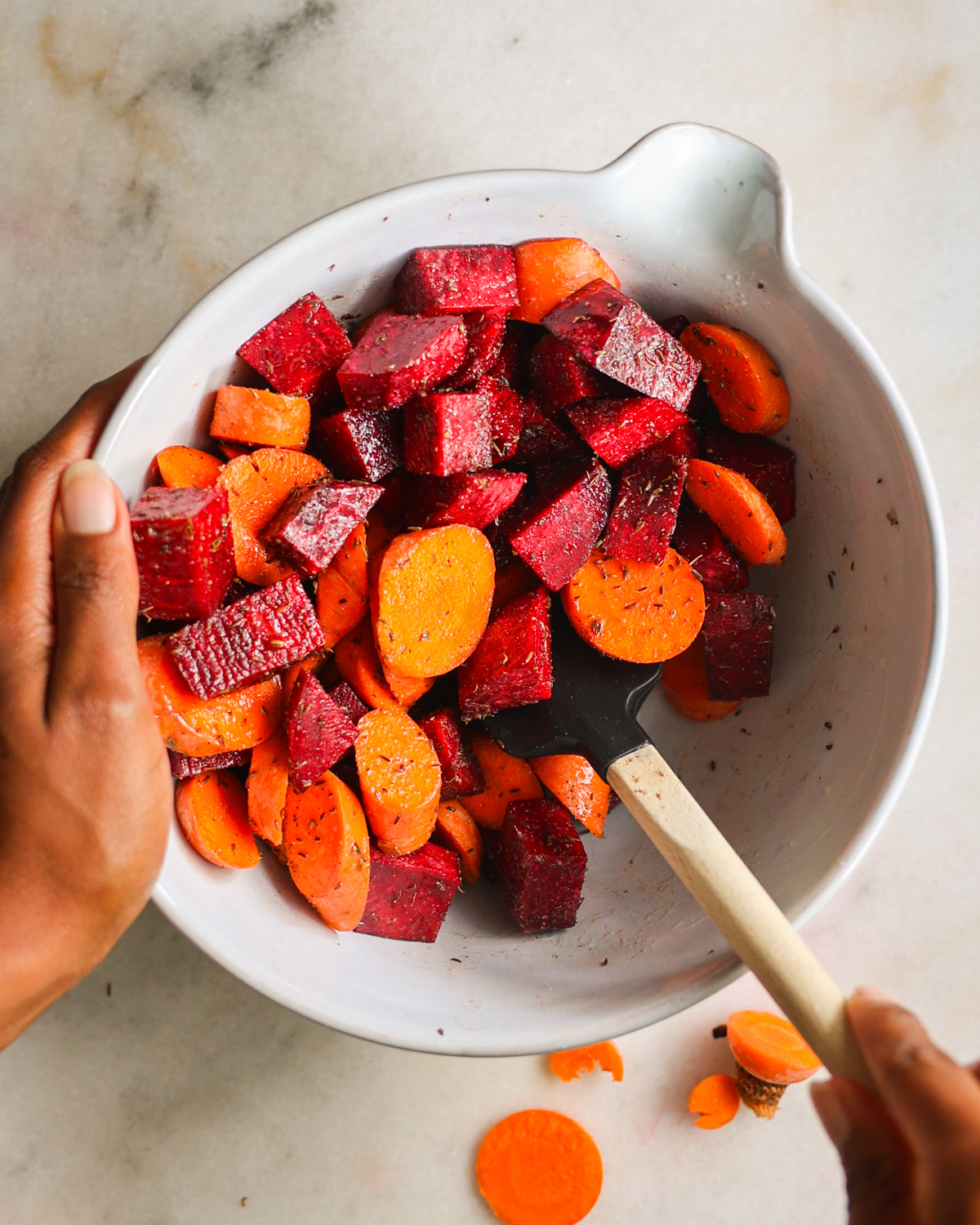 An overhead shot of a brown hand stirring the carrots and beets with seasonings in a mixing bowl.