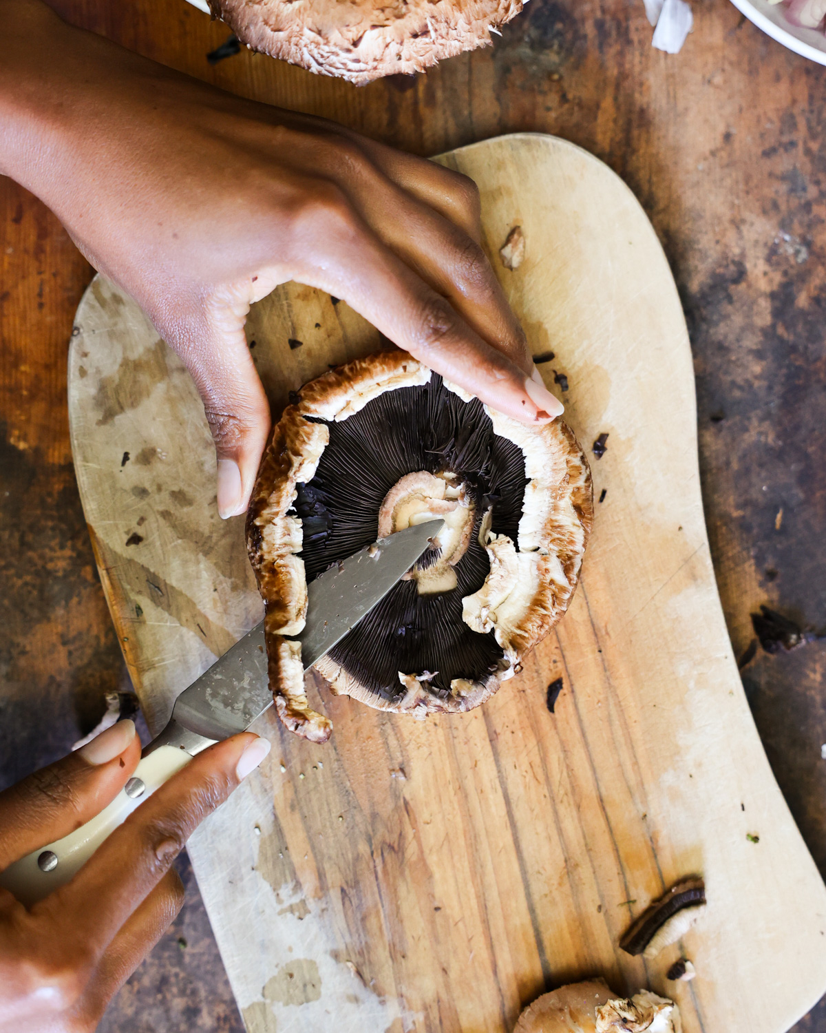 Two brown hands trimming a portobello mushroom on a small cutting board.