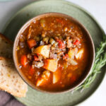 An close-up overhead shot of a small brown cup of the butternut squash and lentil soup with toasted sourdough bread and sprigs of rosemary.