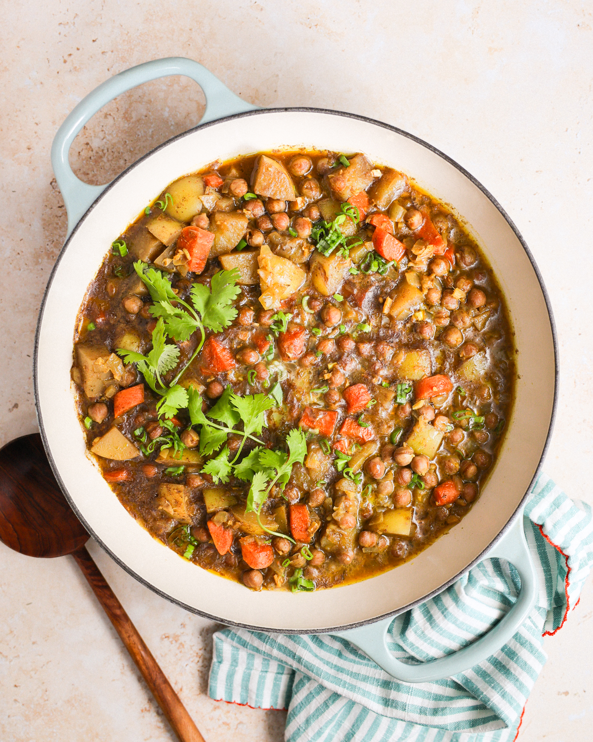 An overhead shot of a white pot of vegan chickpea curry on the counter with a towel and wooden spoon.