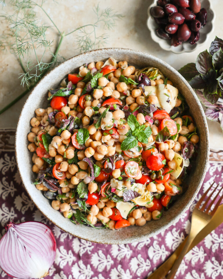 Chickpea salad in a serving bowl framed by olives and herbs.